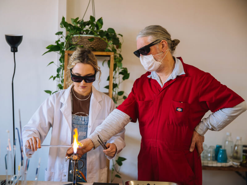 An instructor shows someone how to make hand blown glass in a glass blowing course.